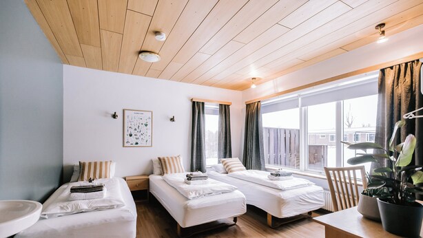 Quarto de hotel com duas camas de solteiro, teto de madeira, uma janela grande com cortinas, uma mesa pequena e um vaso de plantas.