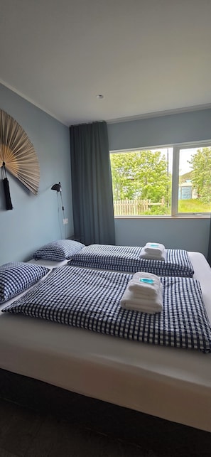 A hotel room with a large bed, a wall fan, a lamp, and a window with a view of trees and a fence. - Eldá apartments (Myvatn)