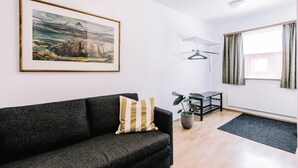 Uma sala de estar moderna com um sofá cinza escuro, uma almofada listrada, um quadro emoldurado de uma paisagem, uma mesinha com uma planta em vaso e uma janela com cortinas.