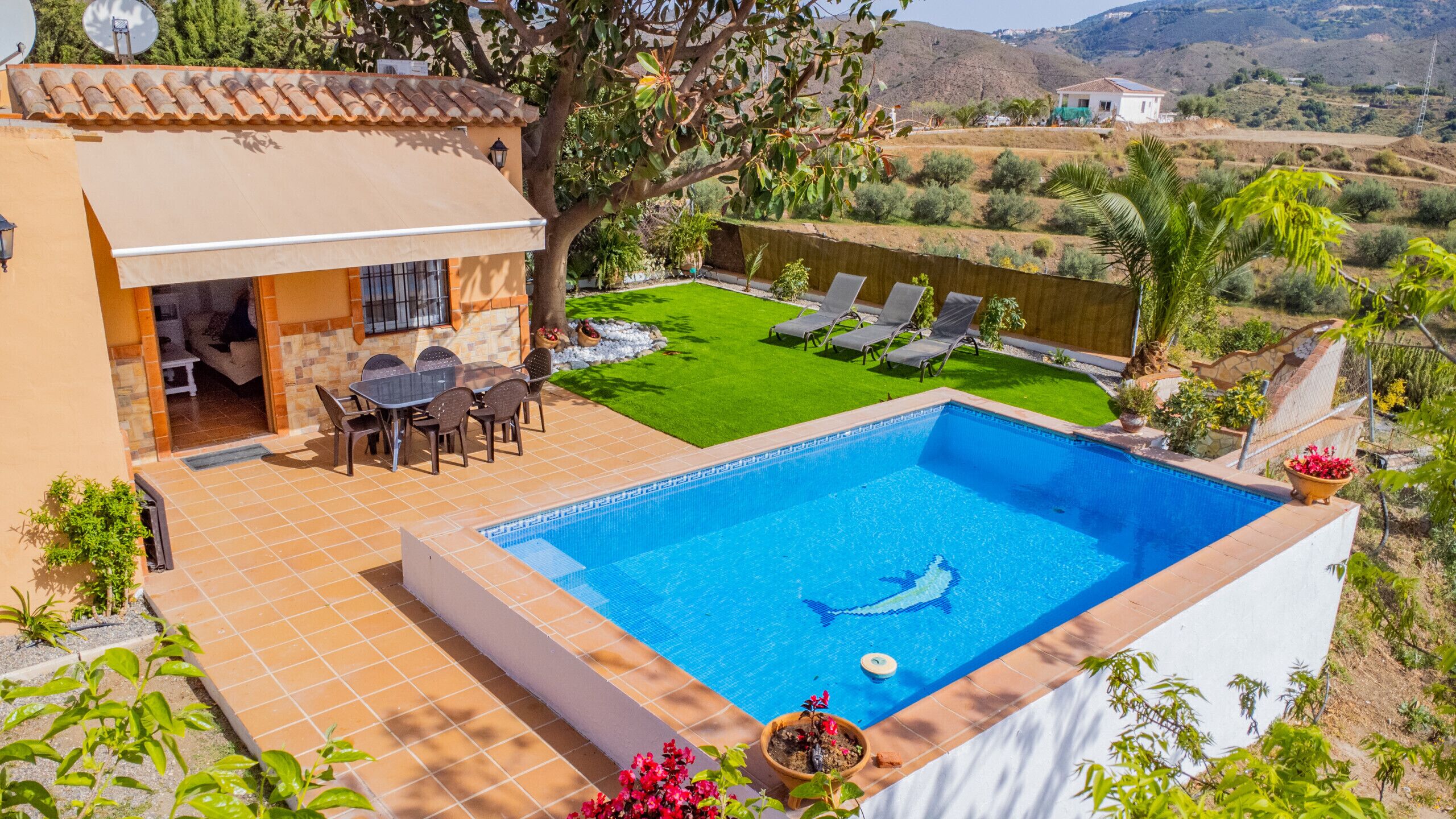 A swimming pool with a dolphin decal, surrounded by a patio with chairs and a table, and a view of a green lawn and distant hills.