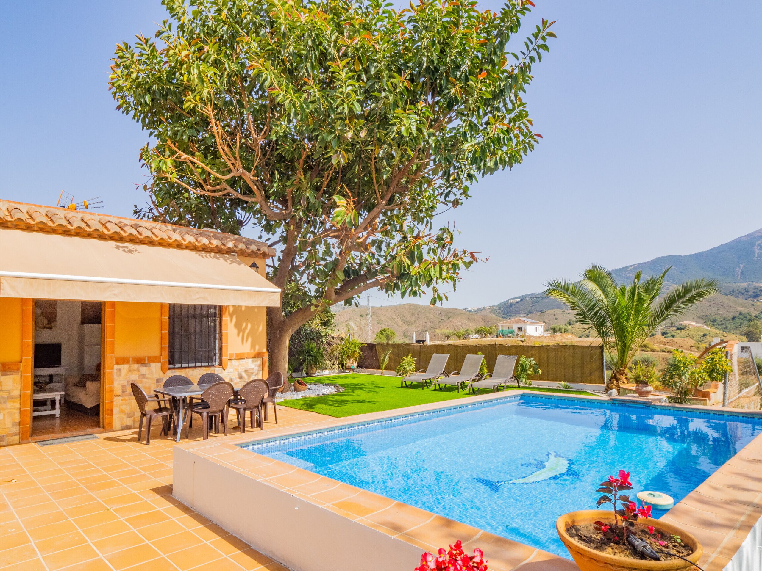 Une piscine avec un espace patio, une table à manger avec des chaises et un grand arbre qui offre de l’ombre.