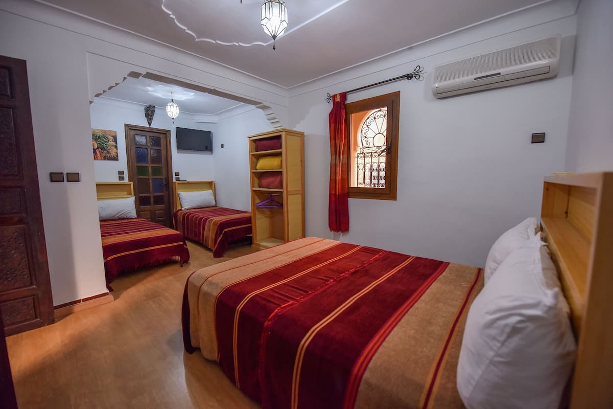A room with three beds, a wooden wardrobe, a built-in shelf, a window with a curtain, and an air conditioning unit.
