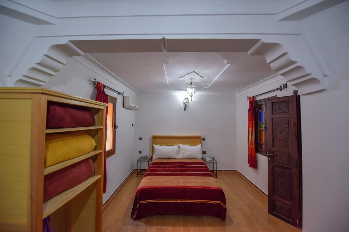 A bedroom with a wooden bed, a shelf with towels, a nightstand, and a door with a decorative panel.
