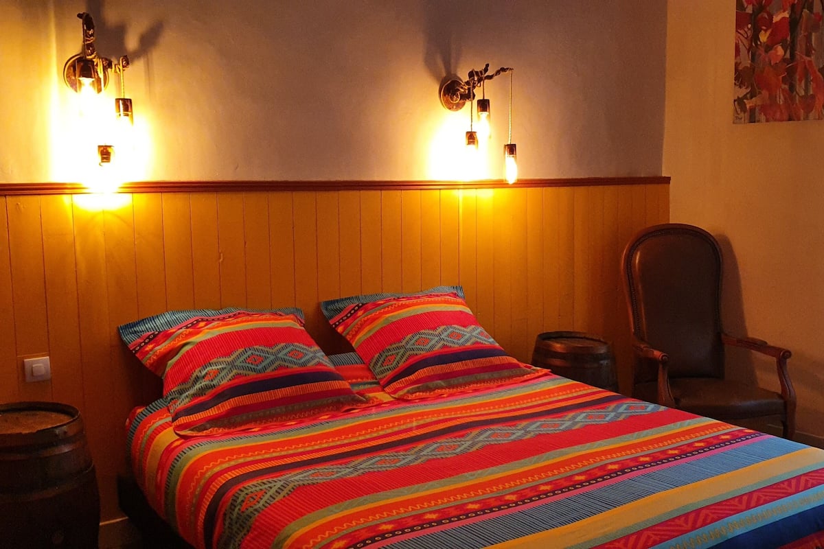 Une pièce avec un mur lambrissé en bois, un lit recouvert d’une couette à rayures colorées, deux lampes fixées au mur, une chaise et une fenêtre ornée de rideaux légers.