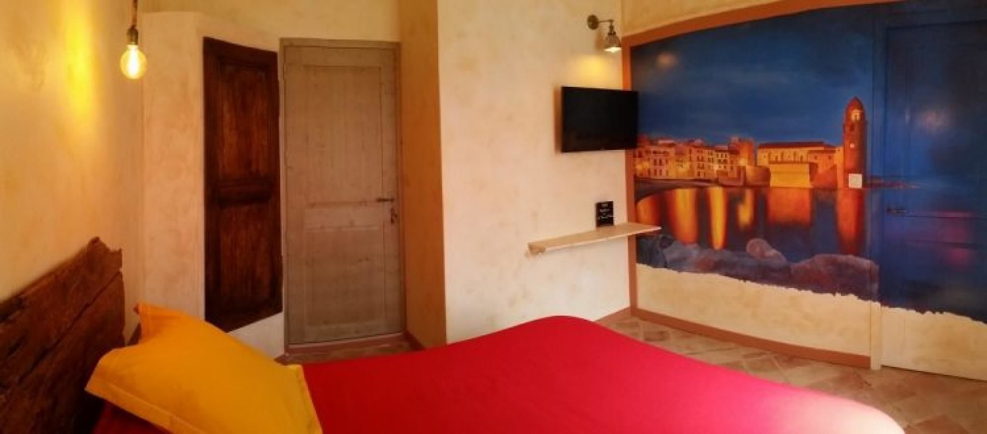 A bedroom with a red bed, a wooden headboard, a door, a wall-mounted TV, and a mural of a cityscape.