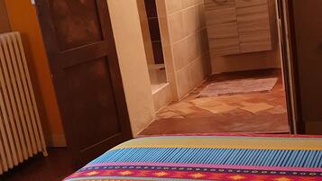 A bedroom with a colorful bedspread, a wooden door, and a view into a bathroom with a tiled shower and a mirror.