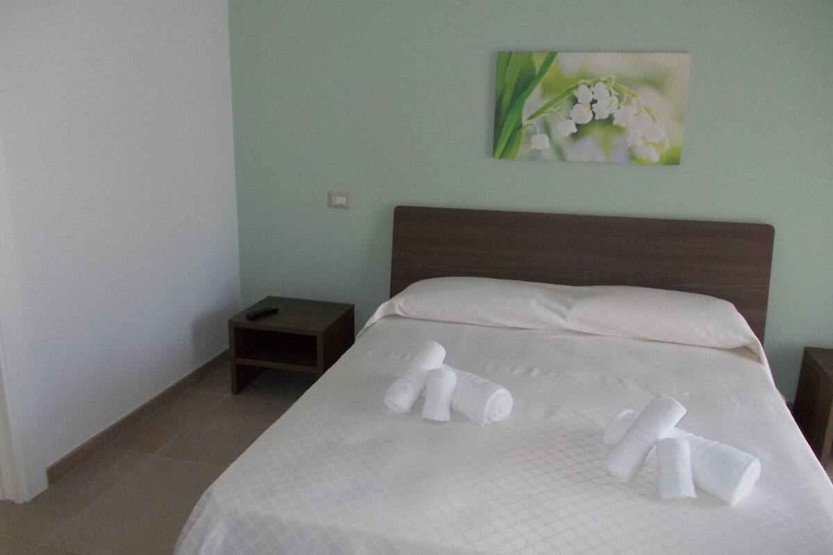 Een netjes opgemaakt bed met witte lakens en opgerolde handdoeken, een klein nachtkastje met een afstandsbediening, en een schilderij van witte bloemen aan de muur.