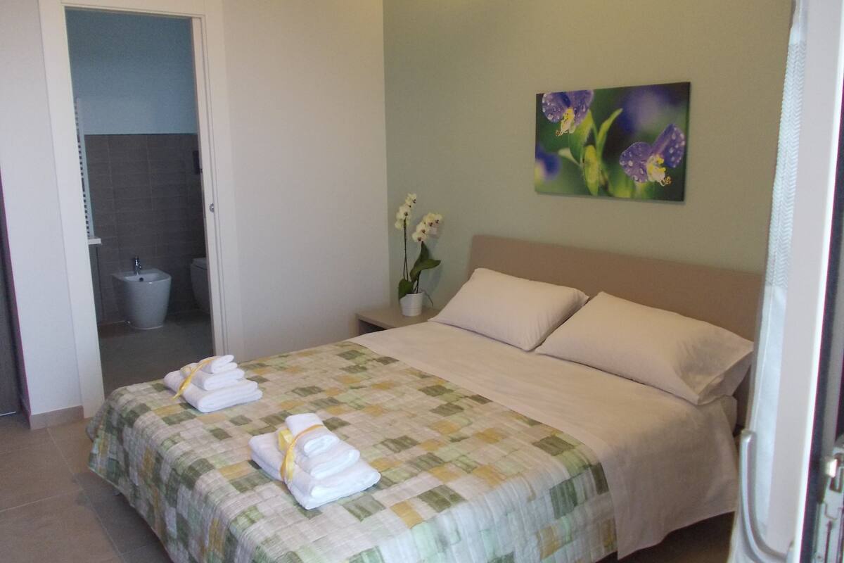Een slaapkamer met een bed, een nachtkastje, een schilderij en een badkamer die door een open deur zichtbaar is.