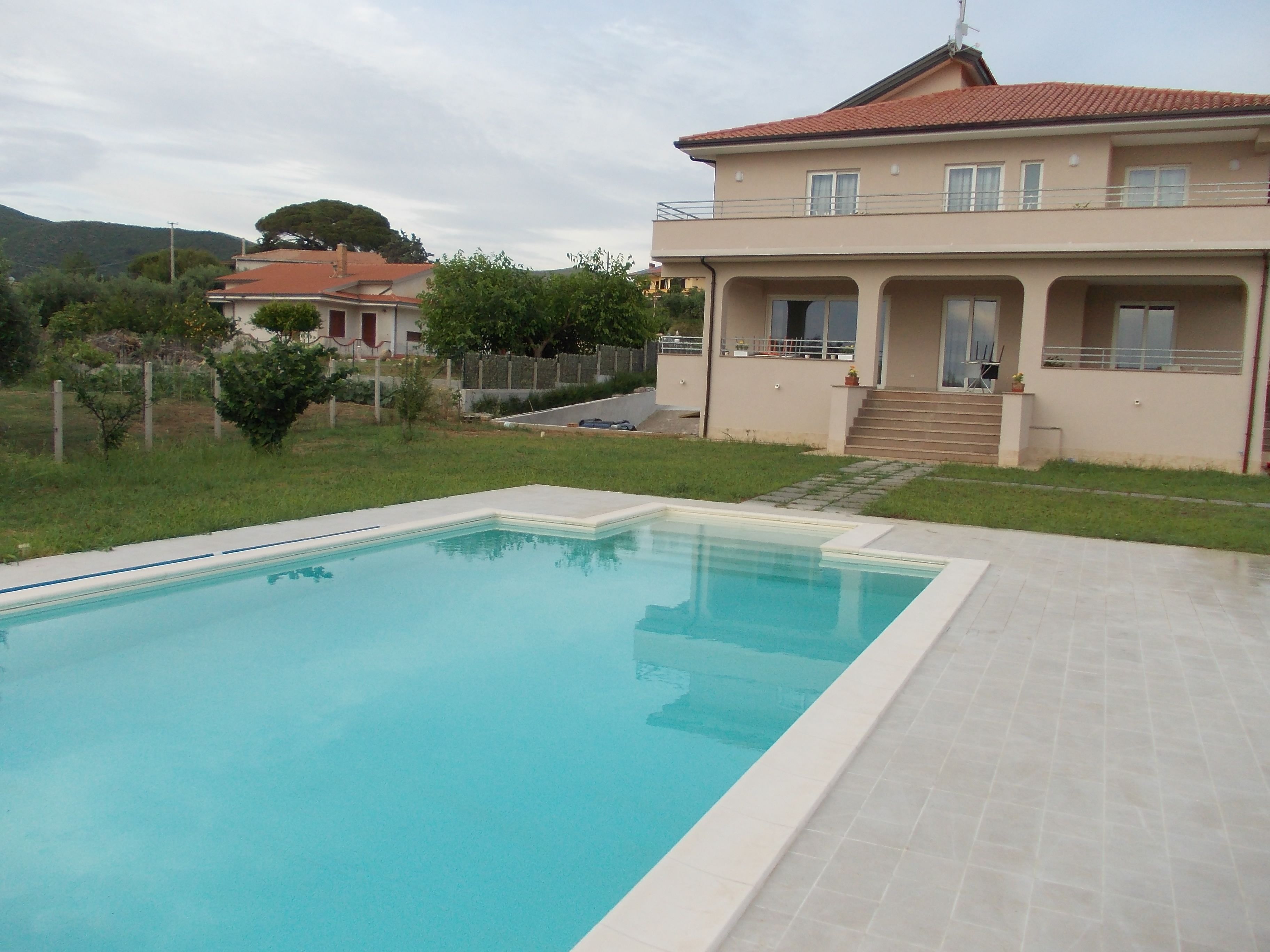 Una piscina con bordo piastrellato, una casa a due piani con un balcone e un prato.