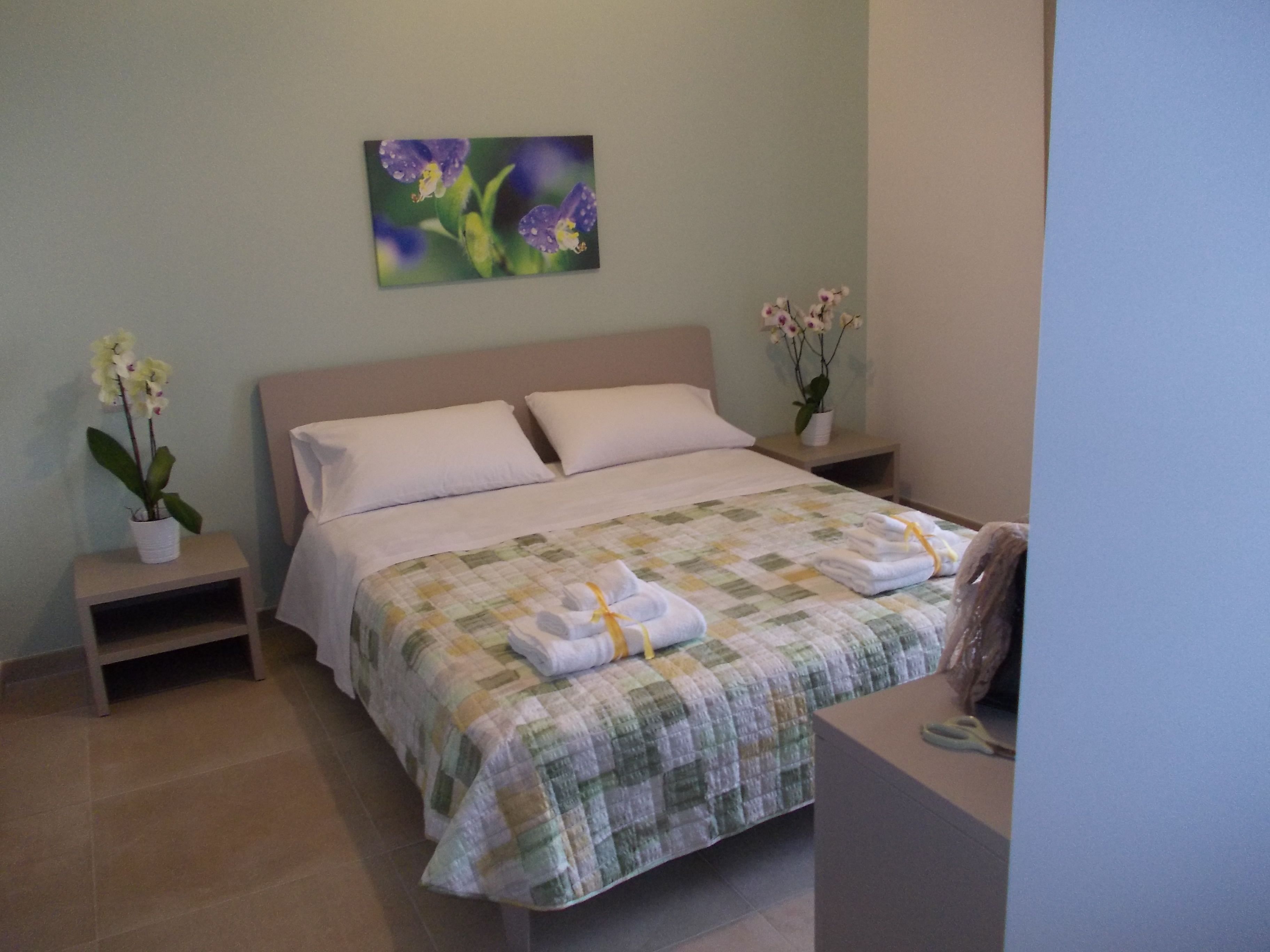A neatly made bed with a quilt, two bedside tables with potted orchids, and a painting of flowers on the wall.