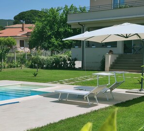 Un espace piscine doté de chaises longues et de parasols, entouré d’une pelouse bien entretenue et d’un bâtiment avec un balcon.
