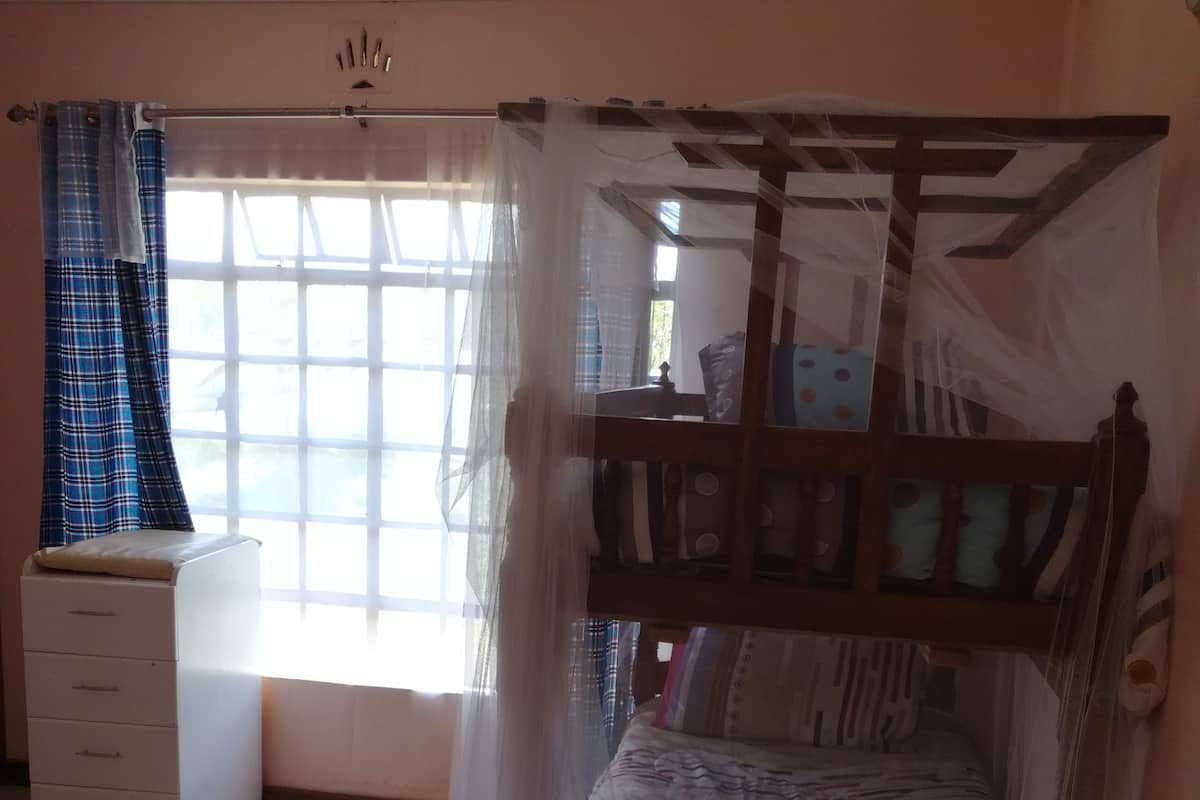 Un dormitorio con una cama nido de madera cubierta por una mosquitera, una mesita de noche, un aparador y una ventana con cortinas.