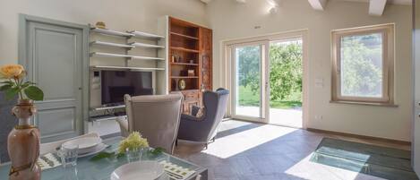 A dining area with a glass table, chairs, a vase with flowers, a television, and a sliding glass door leading to a garden.