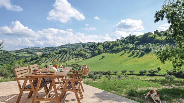 Une table en bois, entourée de chaises, est placée sur une terrasse qui offre une vue imprenable sur un paysage verdoyant, avec des collines ondoyantes et un ciel dégagé.