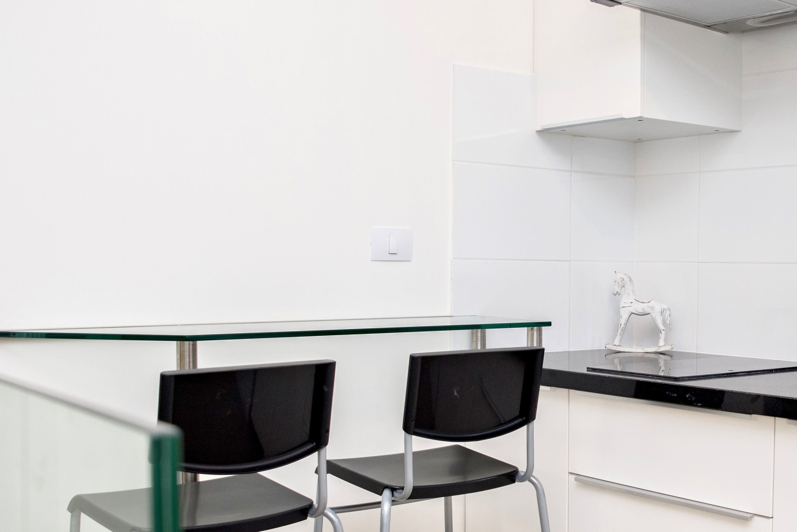 A modern kitchen with two black chairs, a glass-top table, white cabinets, and a white horse-shaped decoration on the countertop.