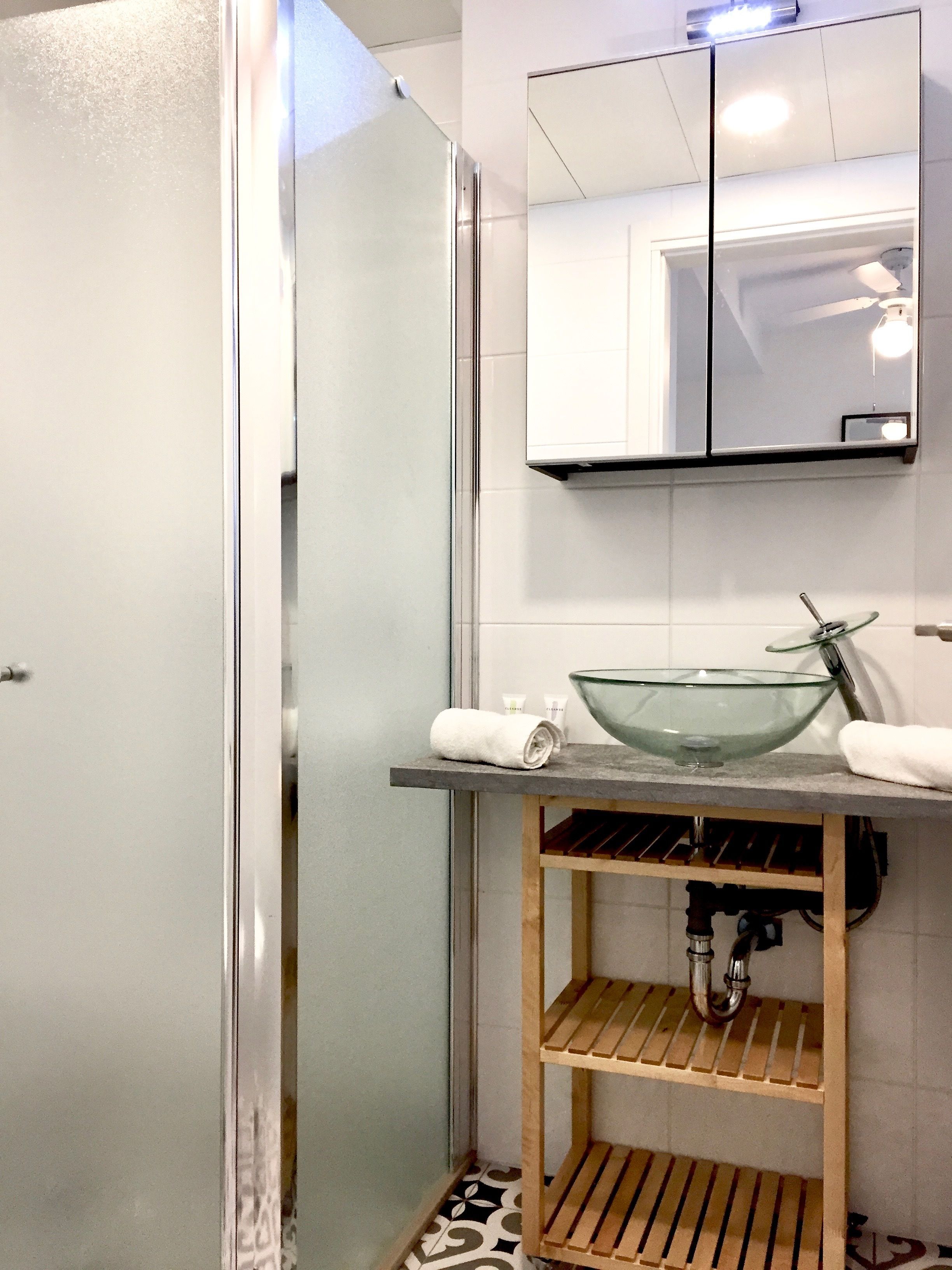 A modern bathroom with a glass bowl sink, wooden shelves, and a glass shower enclosure.