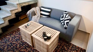 A living room with a grey sofa, two wooden side tables, a patterned rug, and a staircase with a glass railing. - Old Jaffa Designers House (Tel Aviv)