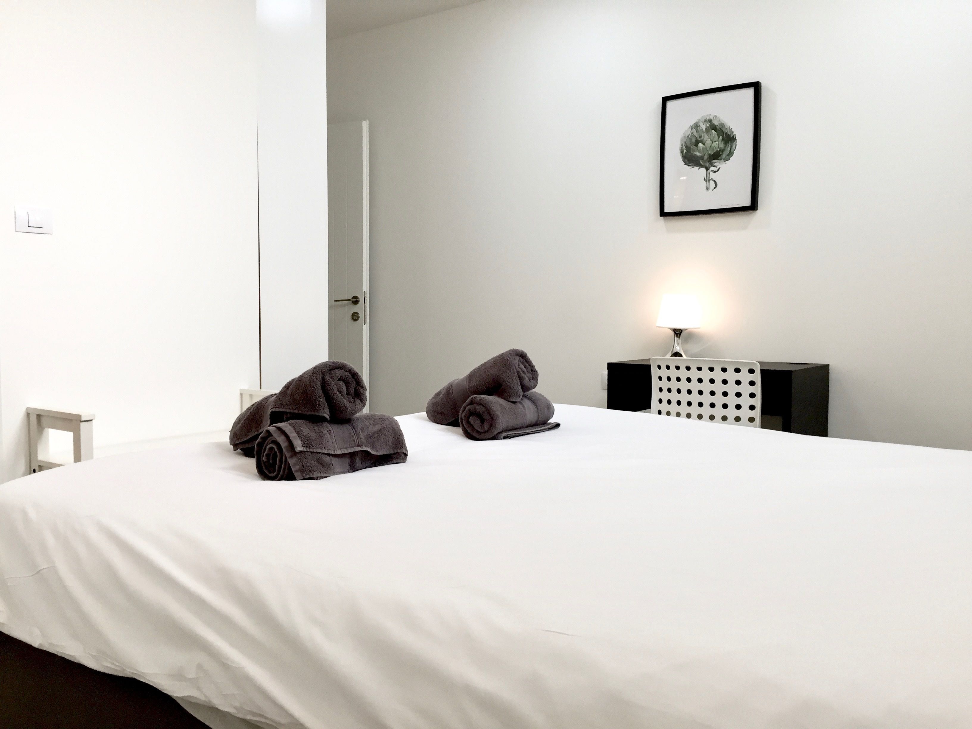 Ein ordentlich bezogenes Bett mit zwei zusammengerollten Handtüchern, ein Nachttisch mit Lampe und ein gerahmtes Kunstwerk an der Wand.