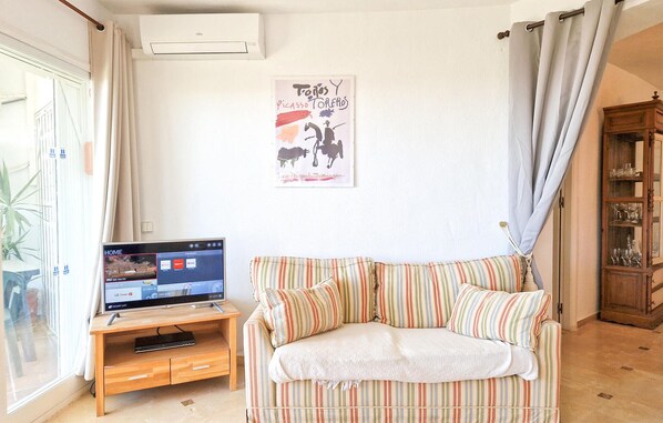 A living room with a striped sofa, a flat-screen TV on a wooden stand, a wall-mounted air conditioner, and a glass door with curtains. - Gorgeous apartment in Mijas with swimming pool (Mijas)