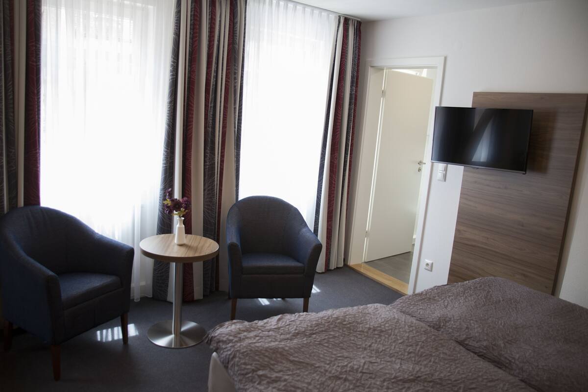 A hotel room with a bed, two armchairs, a small table with a vase, a TV mounted on the wall, and a door leading to another room.