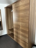 Un armario de madera con dos puertas y un estante, situado en una habitación con alfombra oscura y pared blanca.