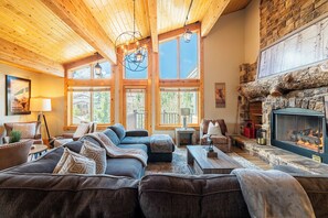 A cozy living room with a fireplace, wooden ceiling, and large windows.
