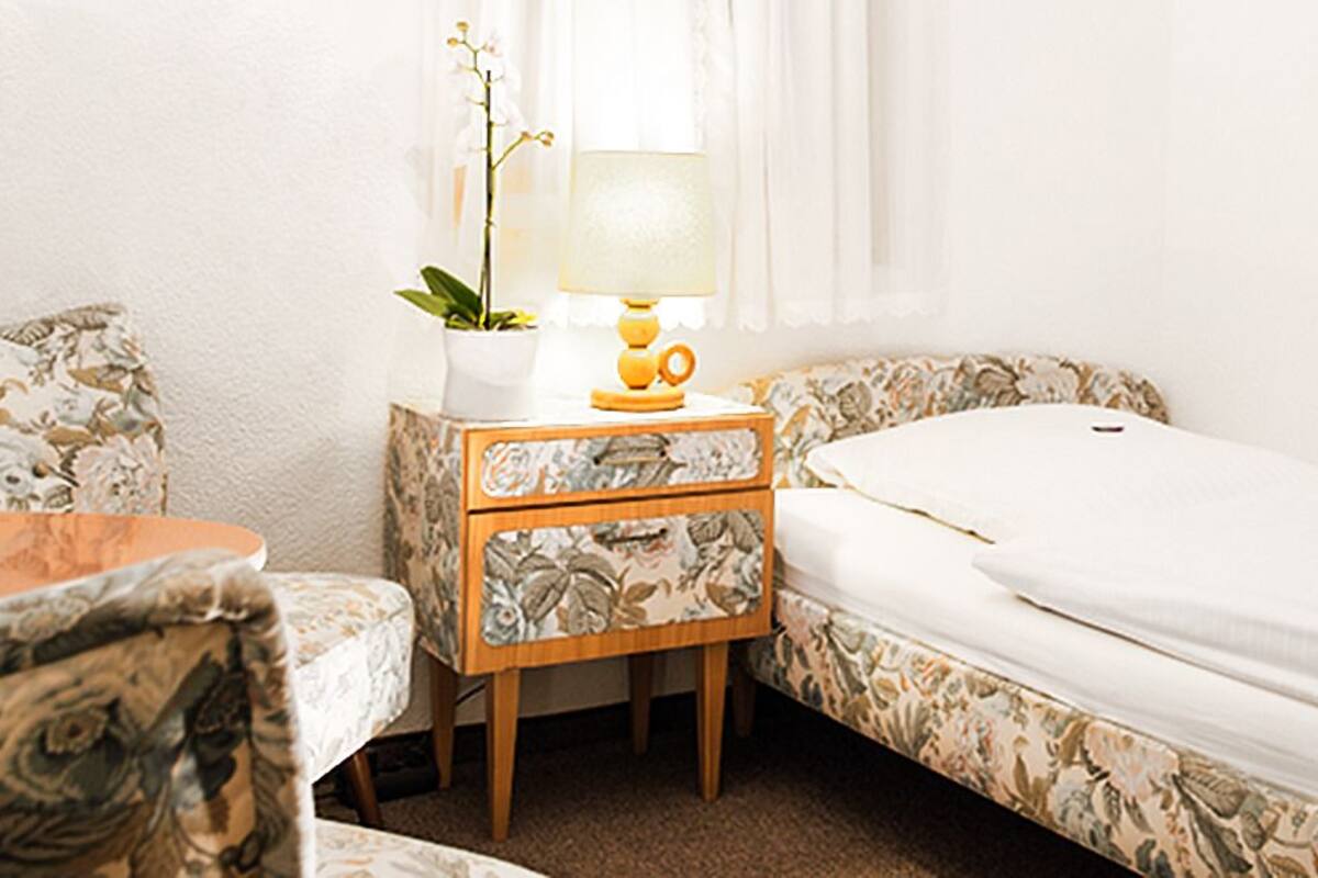 A small, neatly arranged bedroom with a single bed, a bedside table with a lamp, a chair, and a small table with a plant.