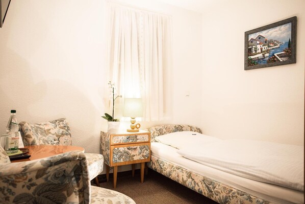 A small, neatly arranged room with a bed, a chair, a nightstand with a lamp, and a painting on the wall.
