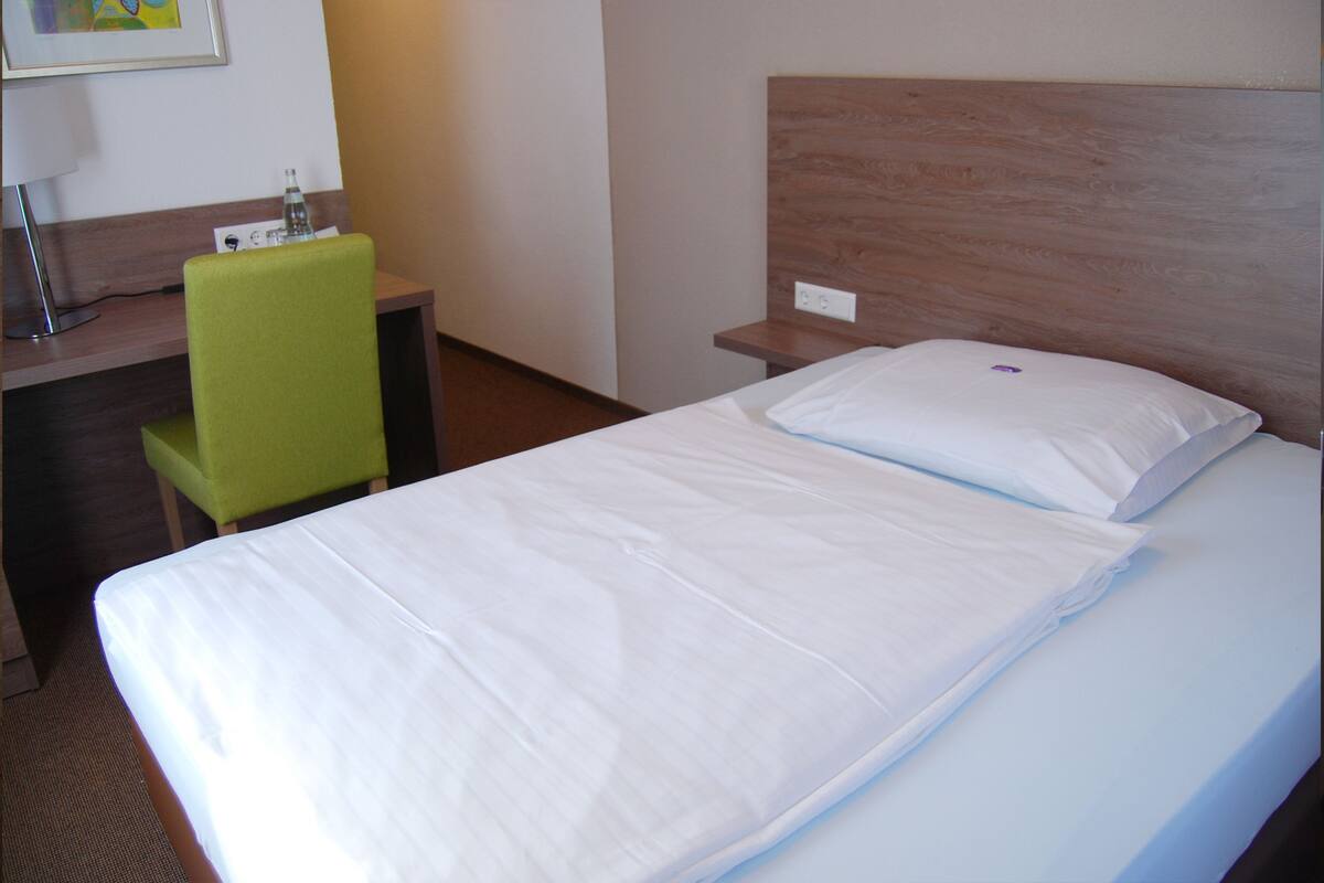 A hotel room with a neatly made bed, a green chair, a desk with a lamp, and a wooden headboard.