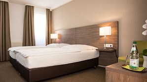 A hotel room with a large bed, a desk with a lamp, a chair, and a bottle on the side table. - Hotel Wagner (Bad Wimpfen)