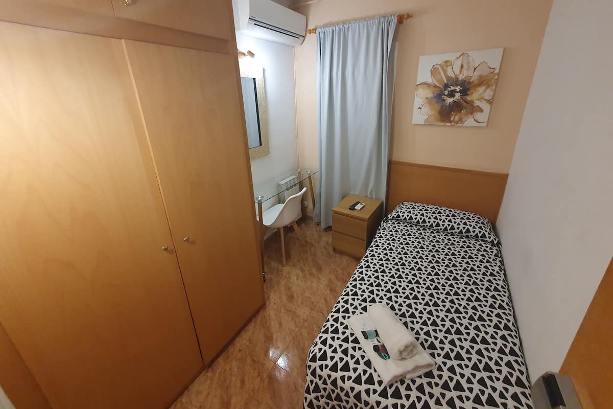 A compact hotel room with a wardrobe, a bedside table with a phone, a bed with a patterned bedspread, and a wall-mounted air conditioner.