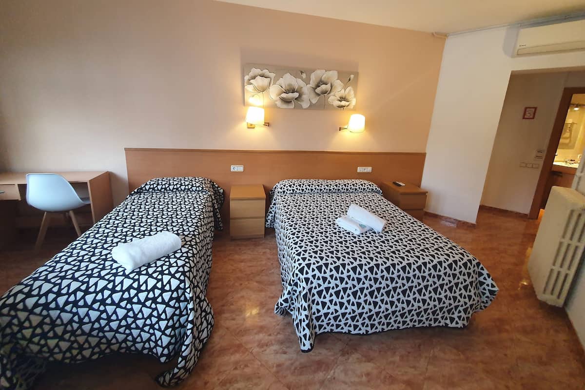 A hotel room with two beds, a desk, and a chair. There are wall-mounted lights and a painting above the beds.