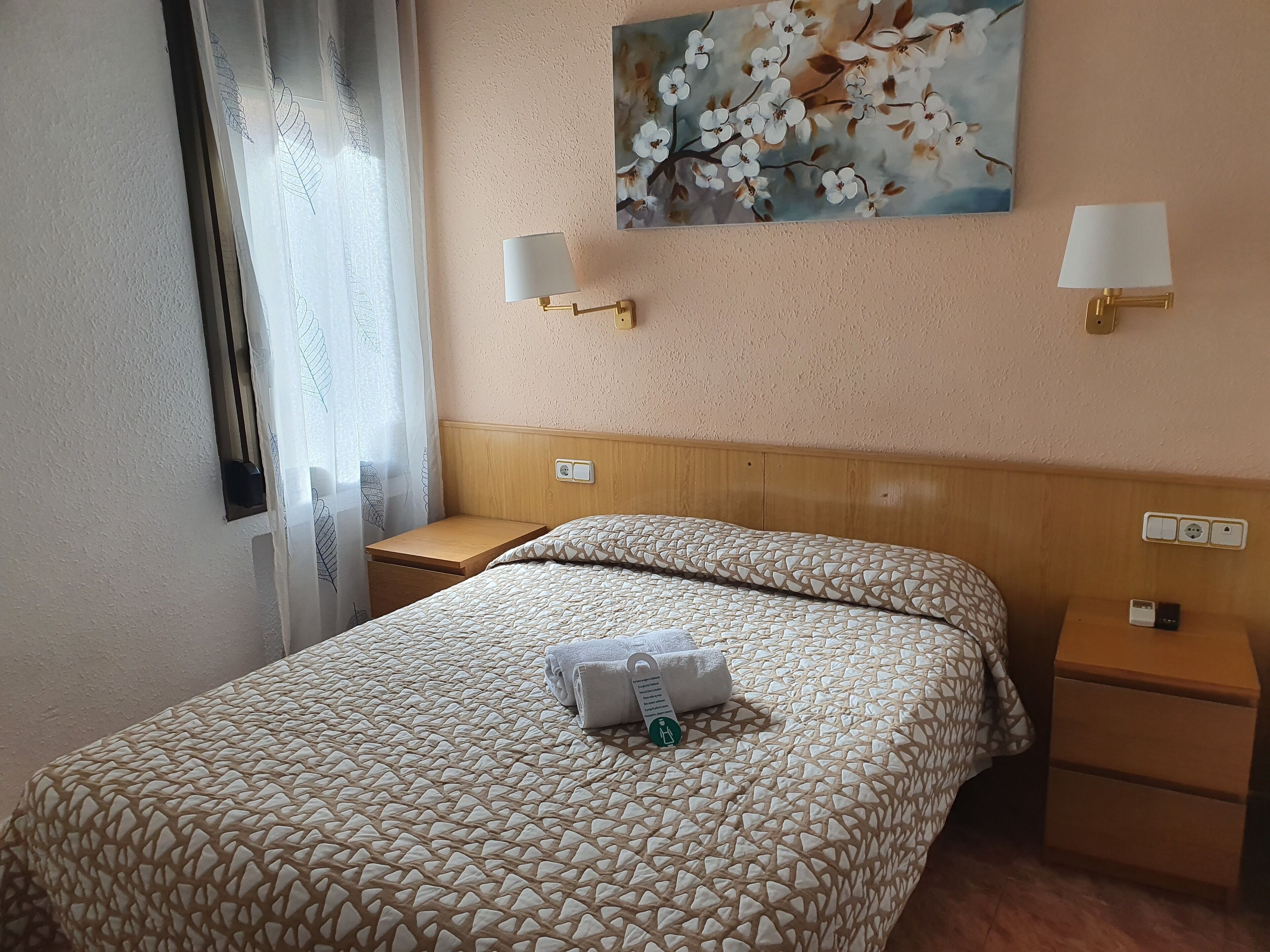 A hotel room with a bed, two bedside tables, a nightstand, a painting on the wall, and a window with curtains.