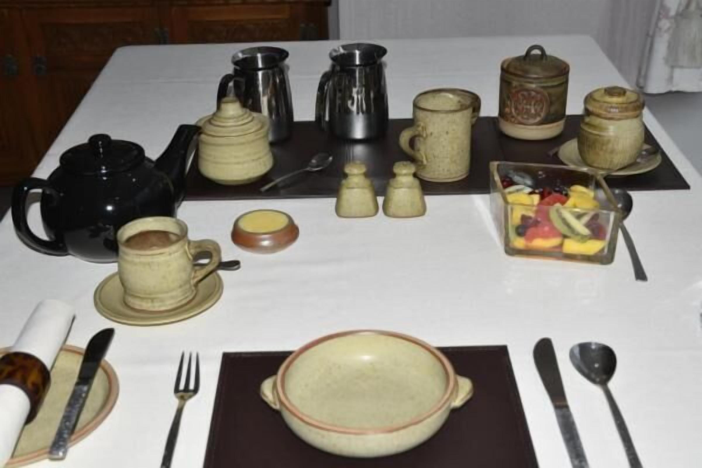 Une table dressée avec différents pots à thé et à café, une théière, des tasses, un saladier rempli de fruits et des couverts.