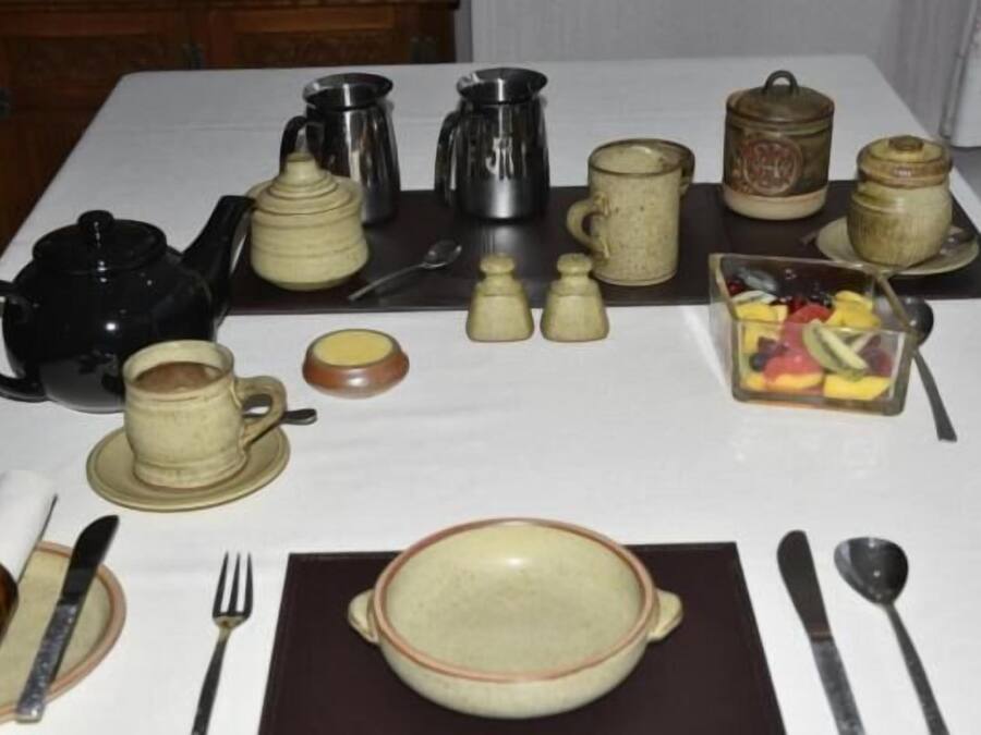 Een gedekte tafel met diverse theepotten en koffiepotten, een theepot, kopjes, een schaal fruit en bestek.