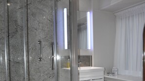 Un baño moderno con ducha envuelta en vidrio, muebles blancos y un ventanal con cortinas.