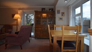 Una sala de estar acogedora con una mesa de comedor de madera, sillas, un sillón con cojines, una lámpara y un aparador con puertas de vidrio.