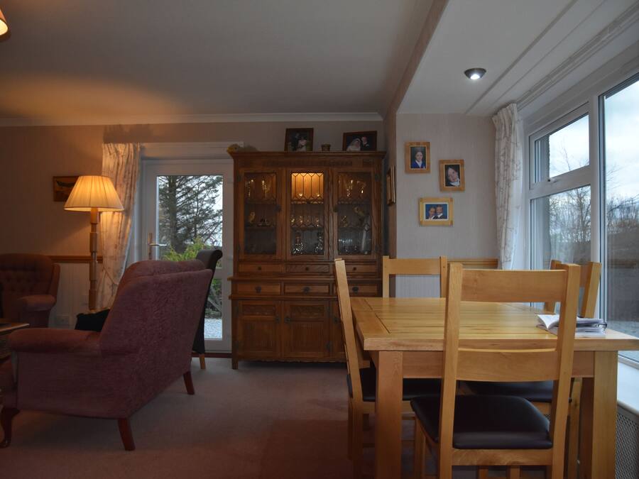 A cozy living room with a wooden dining table, chairs, a cushioned armchair, a lamp, and a cabinet with glass doors.