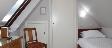 Une chambre avec un plafond en pente, une chaise en bois, un lit avec une literie blanche, une commode et un certificat encadré accroché au mur.
