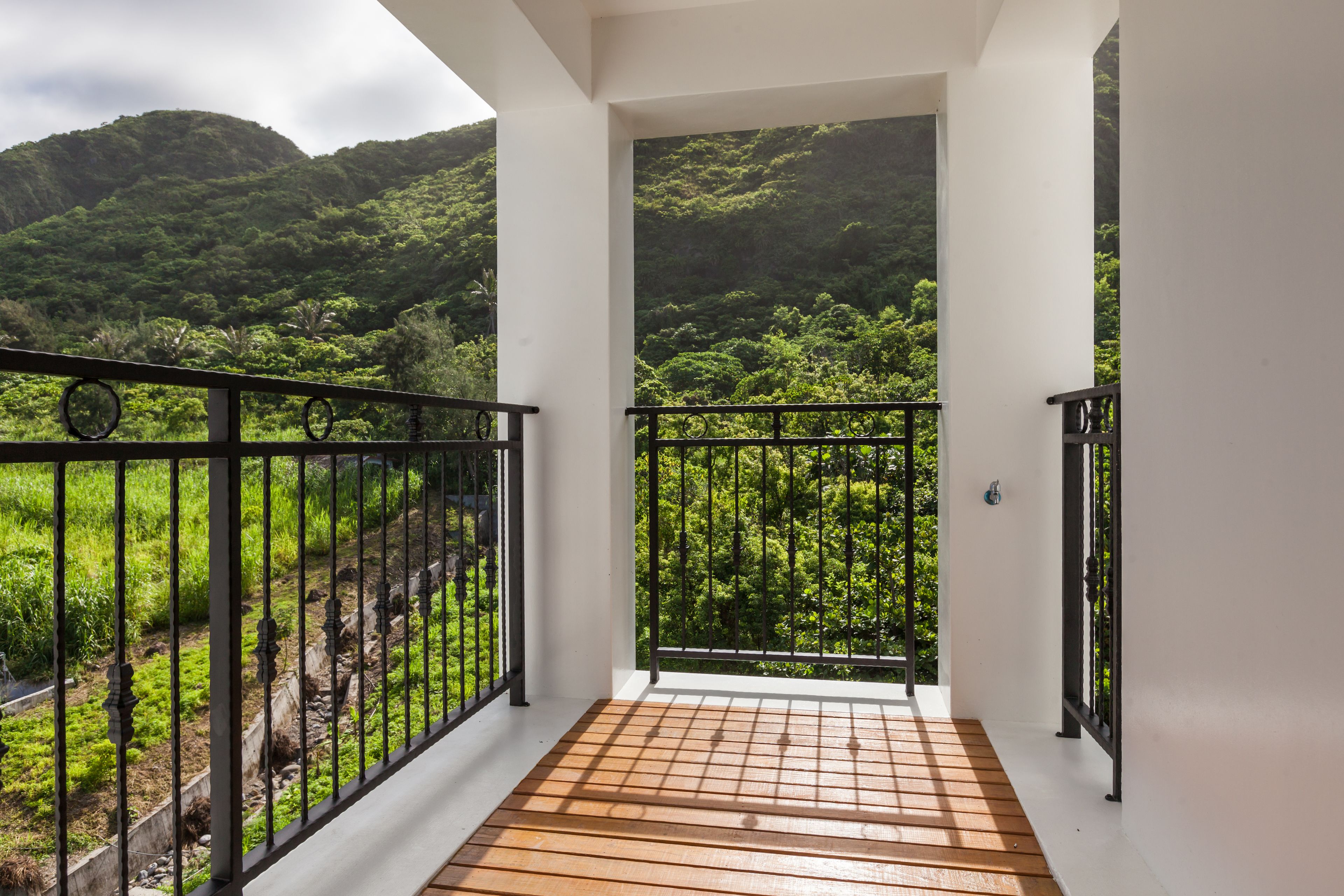 Un balcón con piso de madera y baranda de metal negro, con vistas a un paisaje verde y exuberante con colinas al fondo.