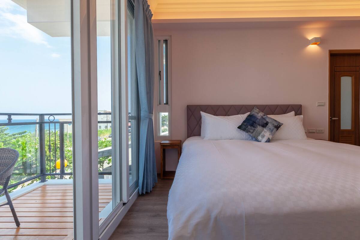 A hotel room with a large bed, a small table, and a chair. There is a balcony with a view of the sea.