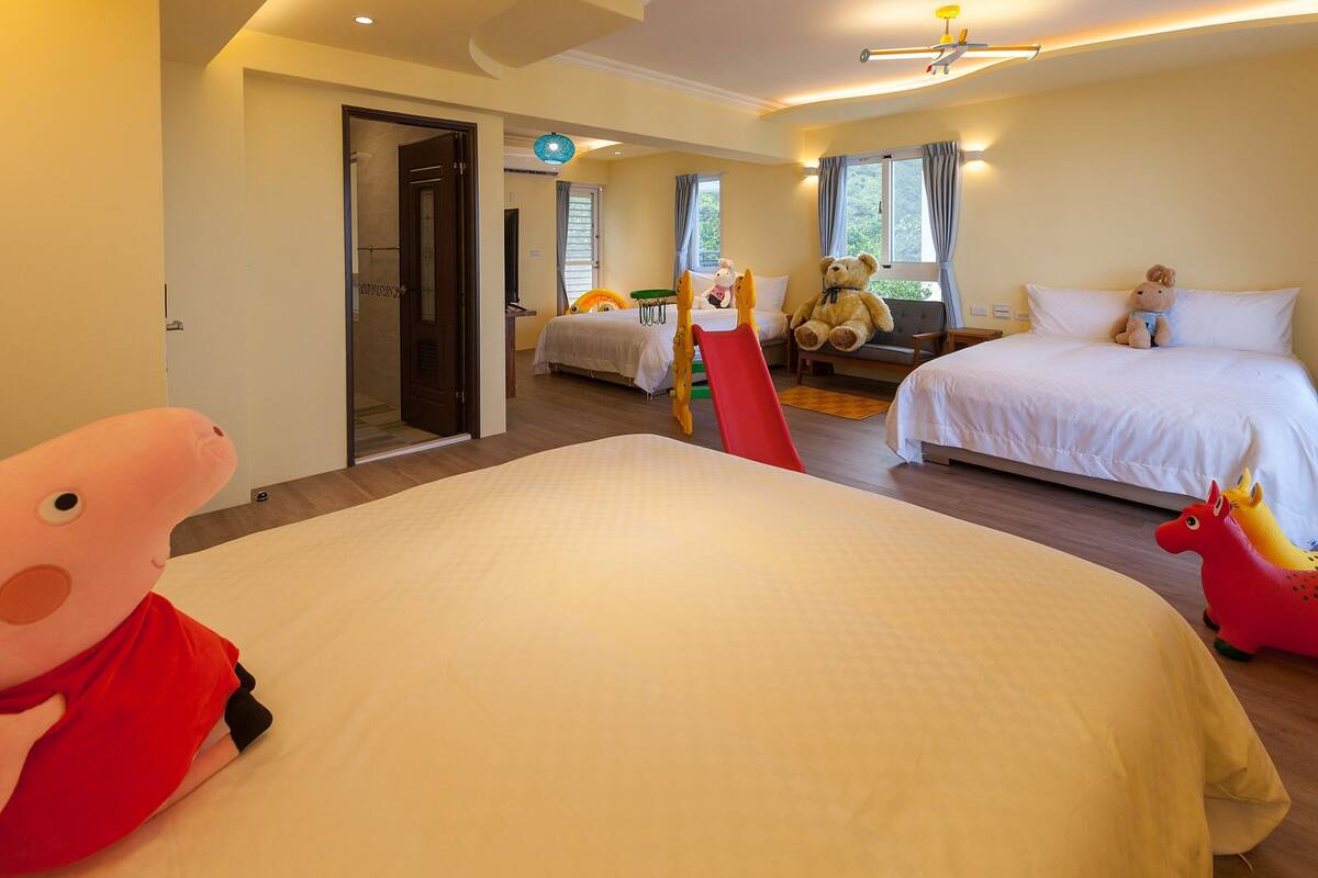 A hotel room with a large bed, a desk, and a chair. There are two windows with curtains, a door, and a small table with a teddy bear.