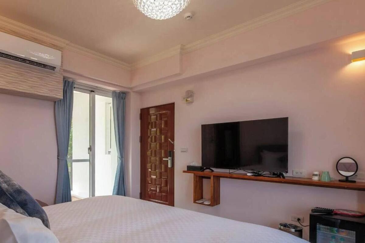 A hotel room with a bed, a TV, an air conditioning unit, a wooden door, and a window with curtains.