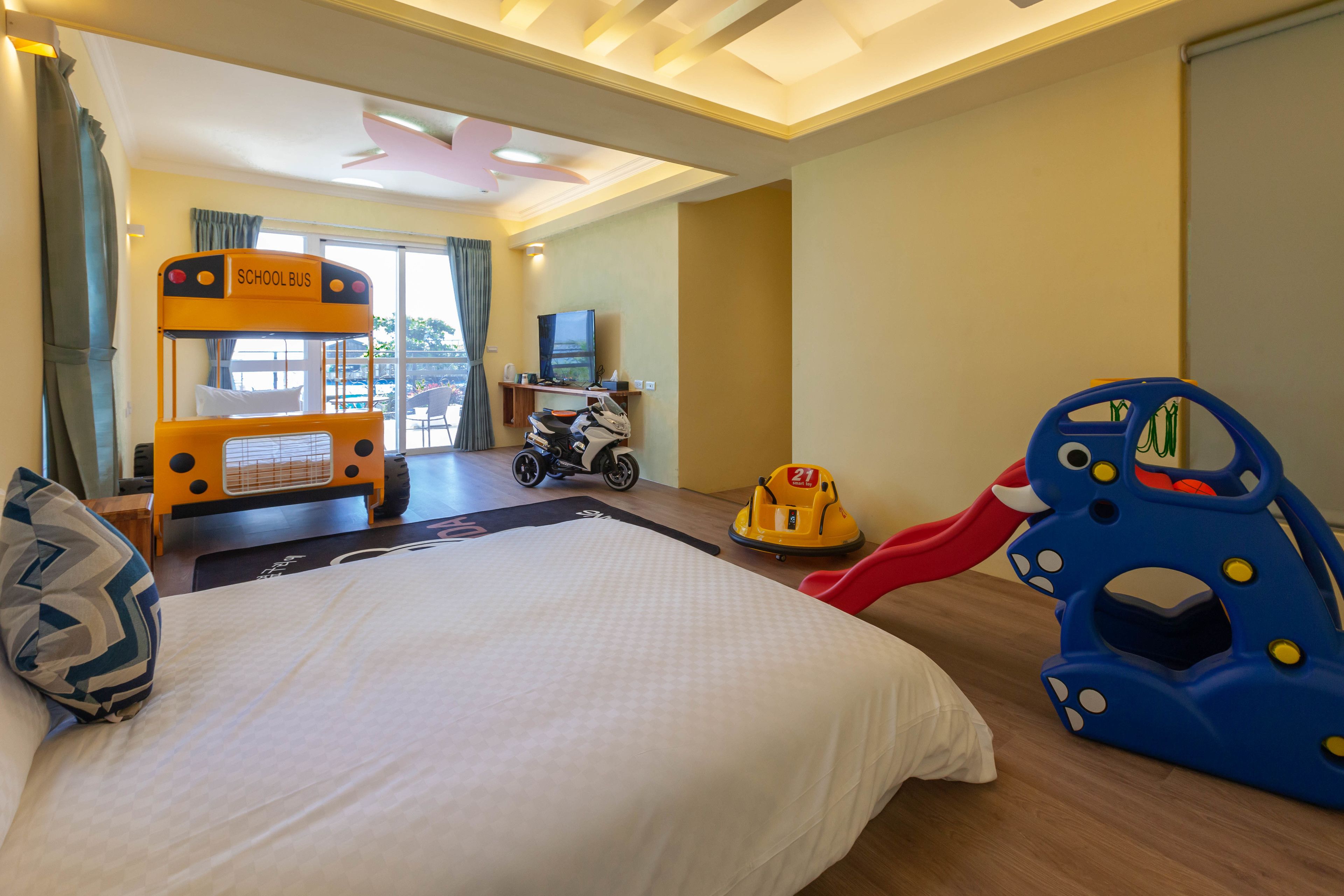 A hotel room with a large bed, a school bus decoration, a toy bus, a toy train, and a television.