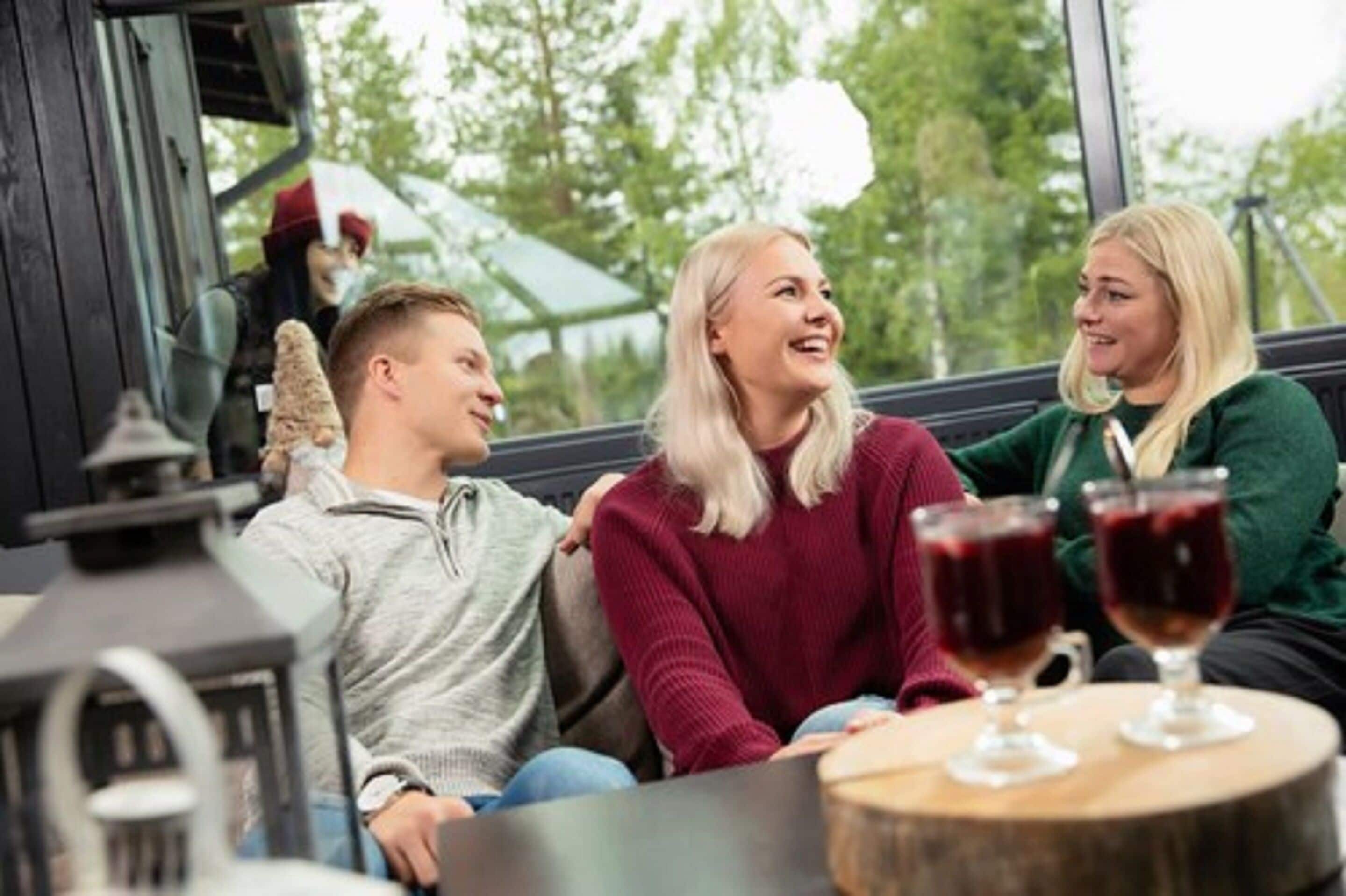 Tres personas sentadas a una mesa con copas de vino tinto, en una habitación con un ventanal y vistas a árboles.