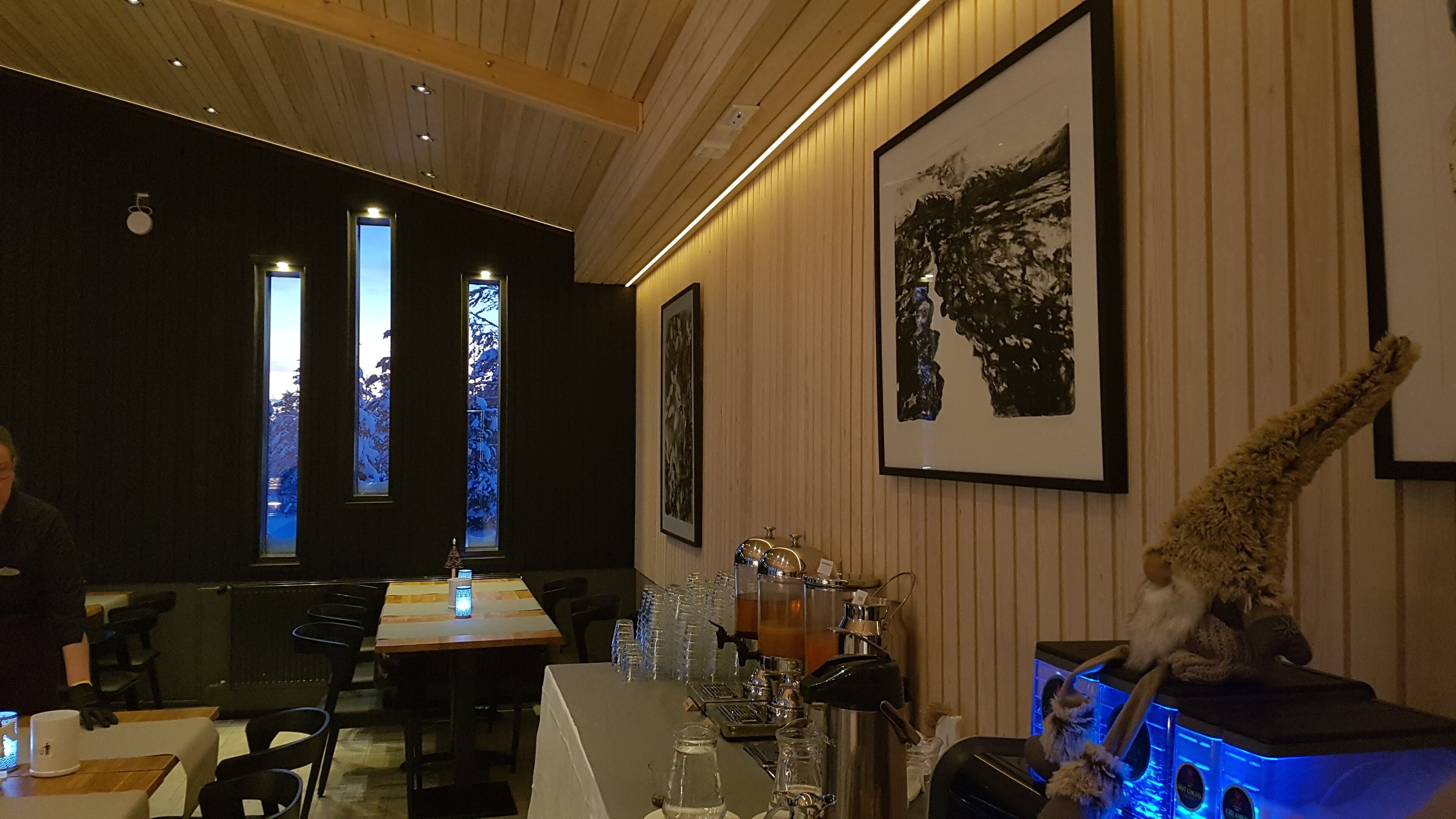 Interior de restaurante con una larga mesa, sillas, una zona de bar con mostrador y una gran obra de arte enmarcada en la pared.