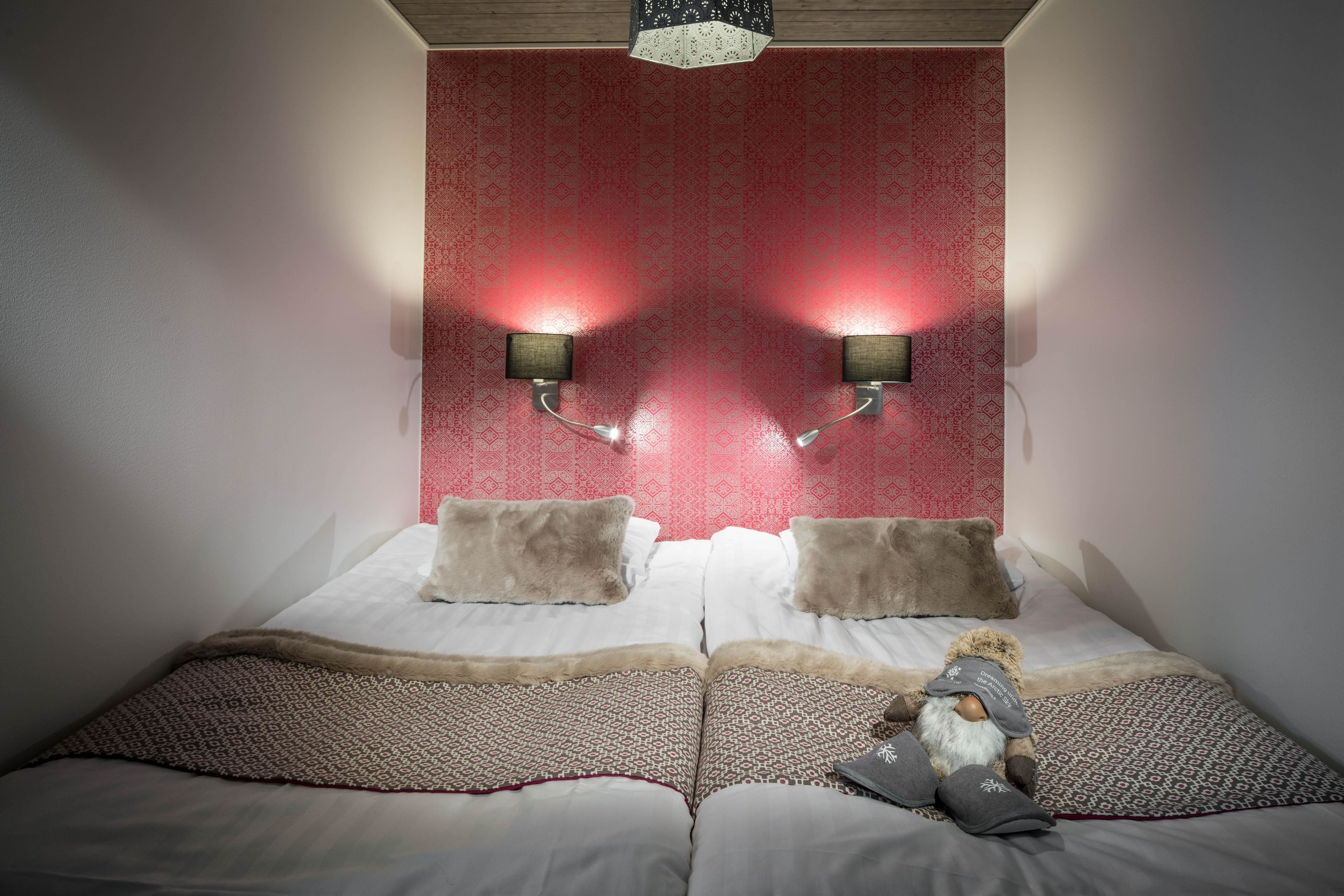 Un dormitorio con papel tapiz rojo estampado, dos lámparas fijadas a la pared, una cama con ropa de cama blanca y dos almohadas, y un pequeño gnomo decorativo sobre la cama.