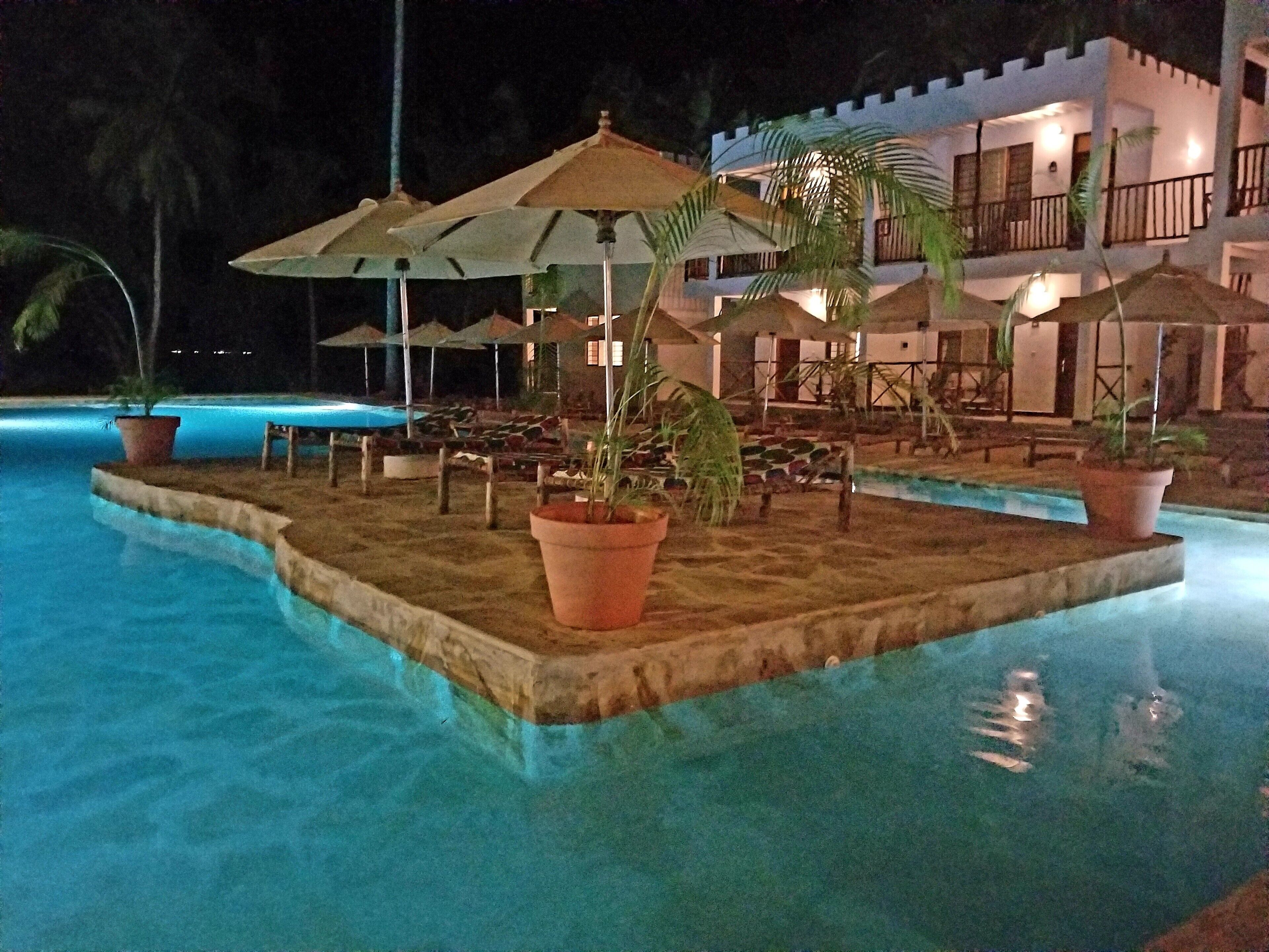 Une zone de piscine éclairée la nuit, avec des parasols, des chaises longues et un bâtiment en arrière-plan.