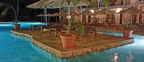 Zona de piscina nocturna con sombrillas, sillas de descanso y un edificio al fondo.