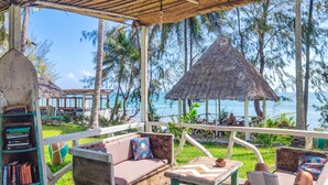 A wooden deck with a thatched-roof gazebo, a cushioned seating area with a book shelf, and a view of the sea and palm trees. - Zanzibar Bay Resort (Marumbi)